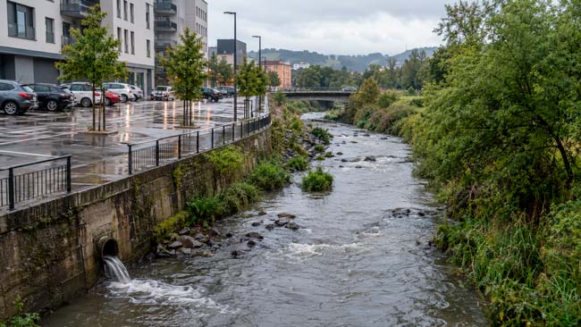 Río urbano junto a calles y aparcamientos impermeables durante una lluvia con escorrentía hacia el cauce